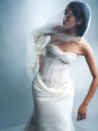 Woman in white strapless gown with a sheer veil poses in a studio with soft, diffused lighting, creating an ethereal and elegant aesthetic.