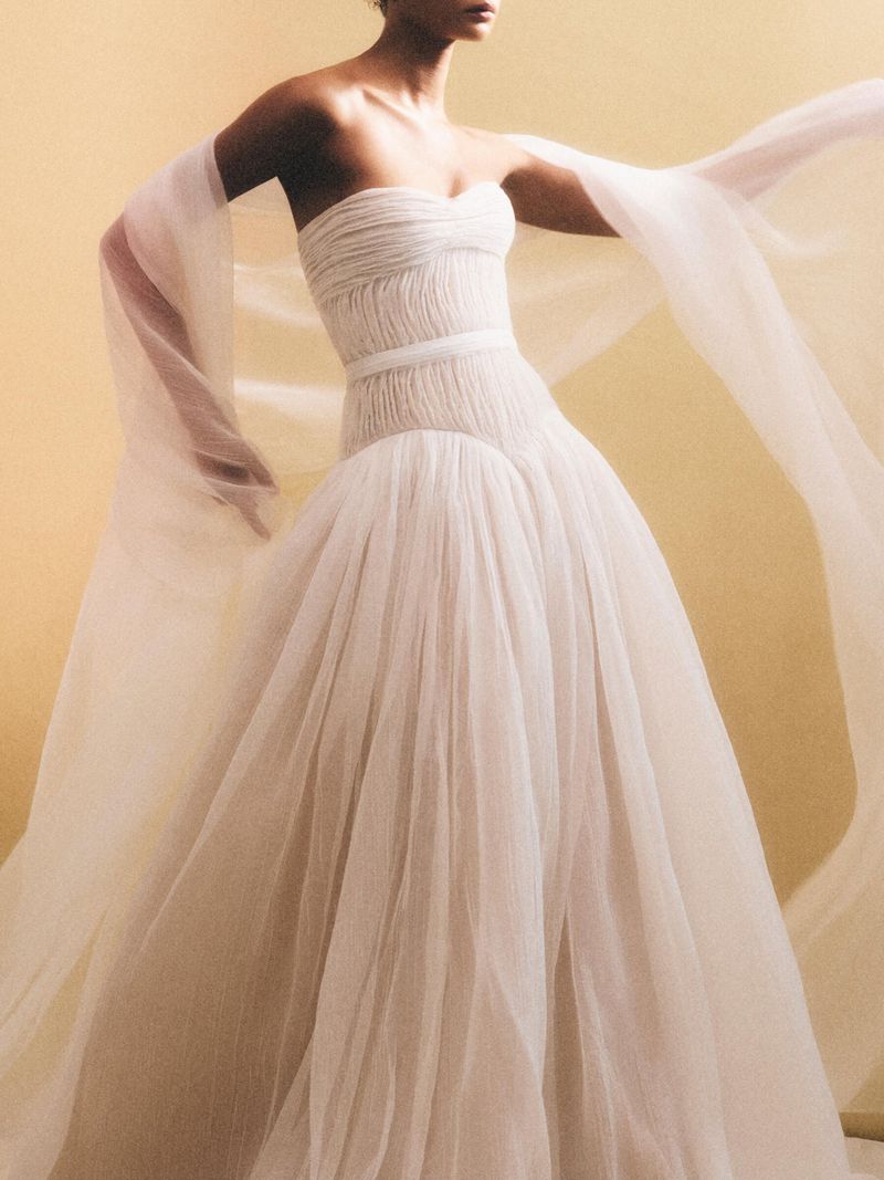 Elegant woman in a white tulle strapless gown with flowing sleeves, posing in a studio with a soft yellow backdrop. 