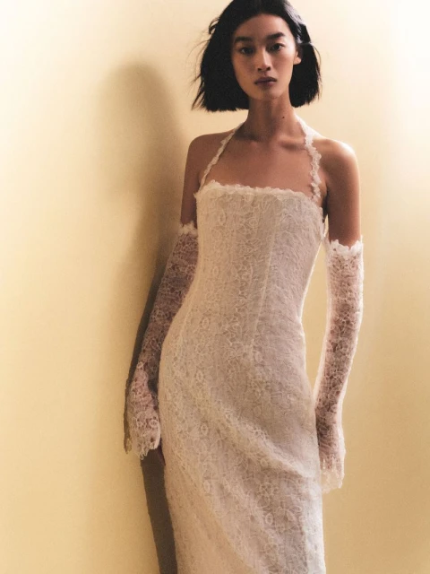 A woman poses gracefully, wearing a form-fitting lace gown with matching arm coverings, set against a neutral backdrop in a portrait shot.