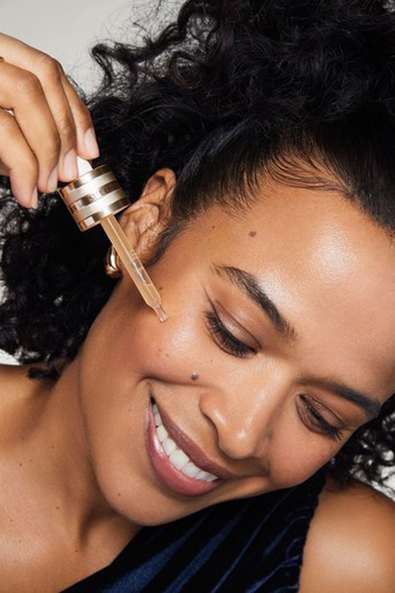 Close-up of a smiling woman applying facial serum to her cheek using a glass dropper, showcasing skincare routine and healthy complexion.