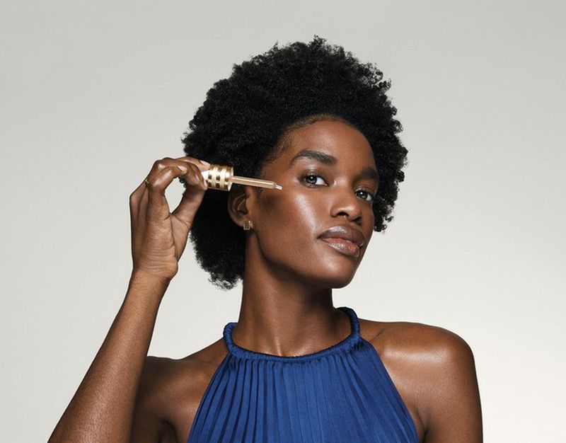 Close-up of Black woman applying facial serum with dropper in studio. Woman with natural hair and blue pleated top.