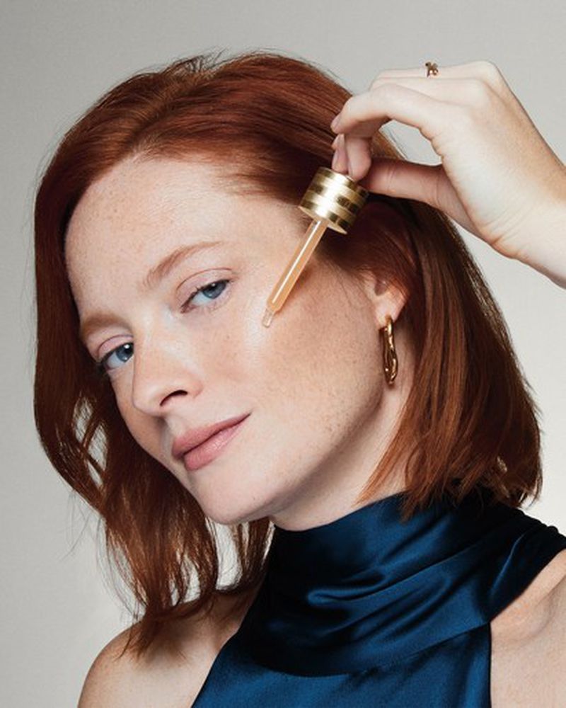 Close-up of a red-haired woman applying serum with a dropper to her face, showcasing beauty and skincare in a bright indoor setting.