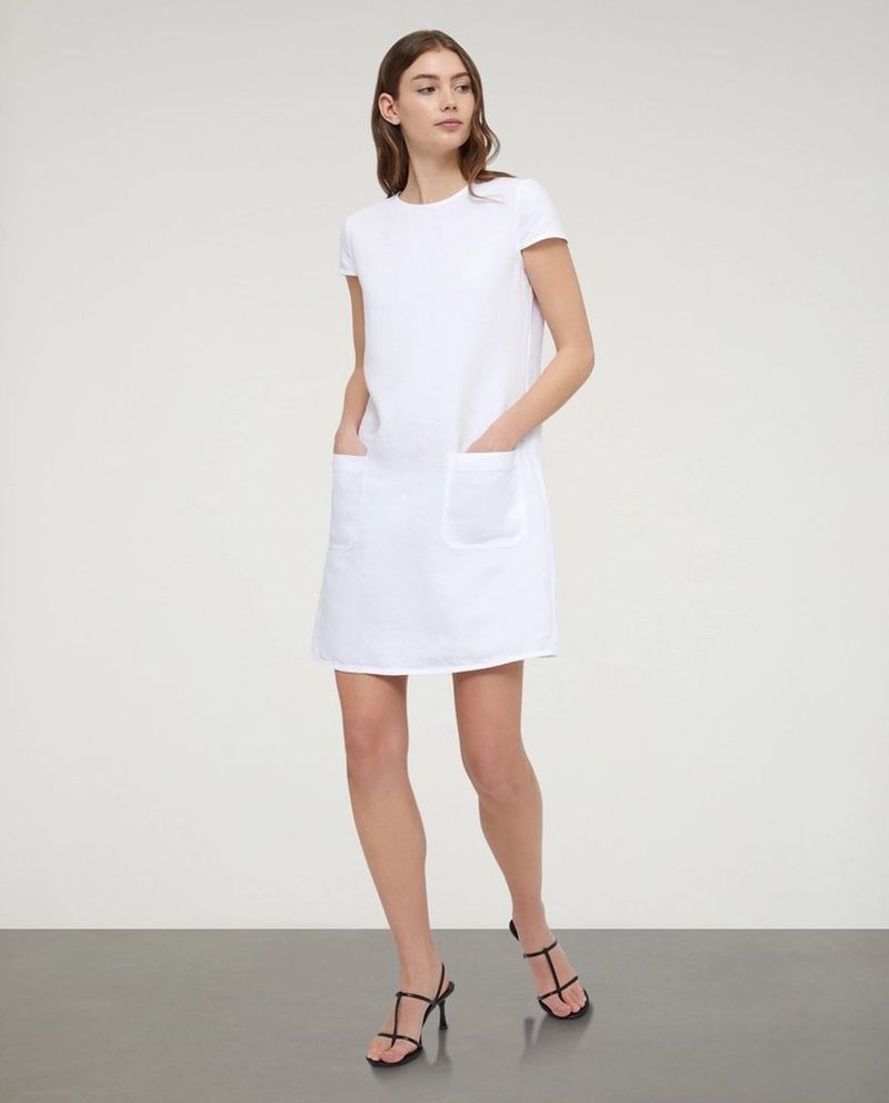 Fashionable adult woman in a short-sleeve white dress and high-heeled sandals strikes a pose in a studio setting with a clean backdrop.
