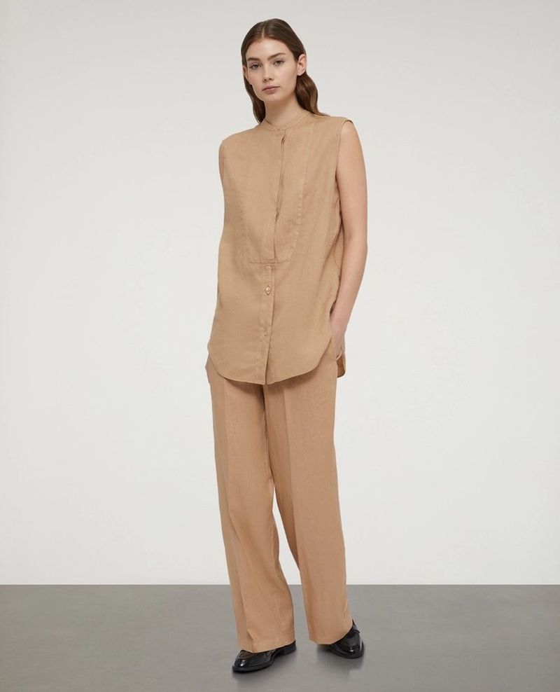 Studio shot of a young adult woman in a tan sleeveless blouse and tan wide-leg pants, posing against a pale gray background.
