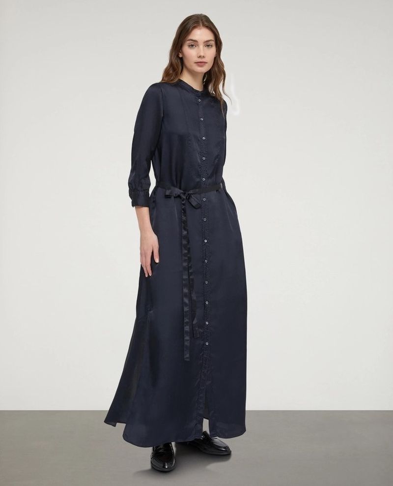 A woman in a navy, full-length button-down dress with a belt stands in a studio with a neutral background, looking directly at the camera.