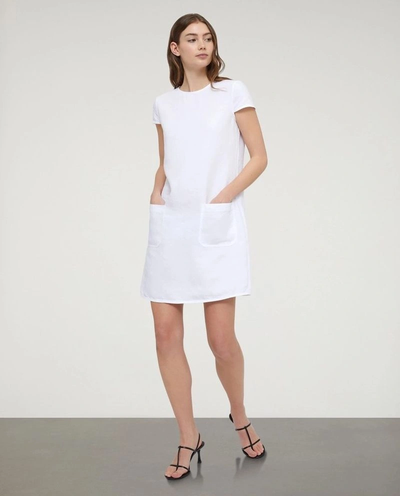 Model standing against a neutral studio backdrop wearing a white dress with pockets and black strappy sandals.