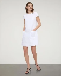 Model standing against a neutral studio backdrop wearing a white dress with pockets and black strappy sandals.