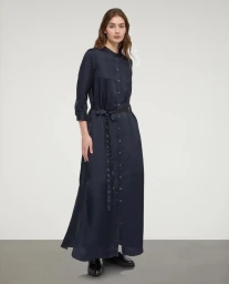 Model standing against a neutral studio backdrop wearing a long dark button-down maxi shirt dress with a matching waist tie.