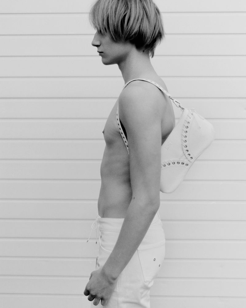 Side profile of an adult male wearing white jeans and a bag in front of a white horizontal paneled wall in black and white.