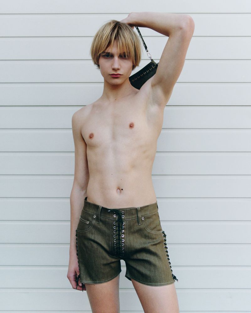 A fair-skinned shirtless person with blonde hair holds a black purse with a white horizontal slat wall as a backdrop.