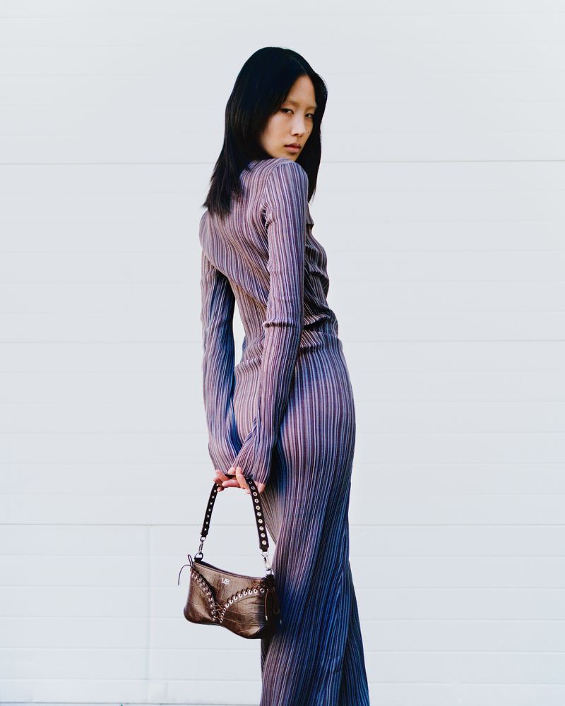 Attractive Asian woman in purple pleated dress and small brown purse poses against a white wall, looking over her shoulder.