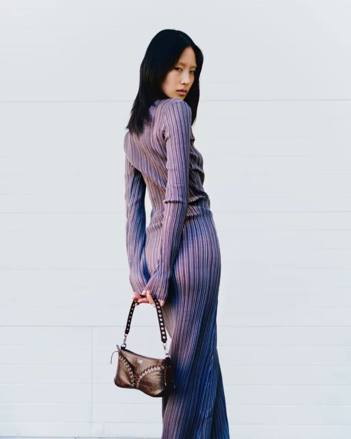 Attractive Asian woman in purple pleated dress and small brown purse poses against a white wall, looking over her shoulder.