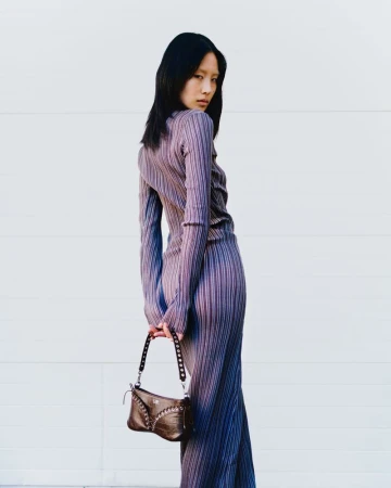 Attractive Asian woman in purple pleated dress and small brown purse poses against a white wall, looking over her shoulder.