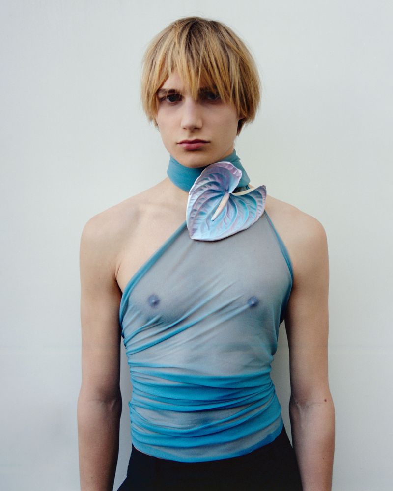 Person poses in a blue, sheer top with a large flower on the shoulder against a plain white background in a studio shot. Fashion forward.