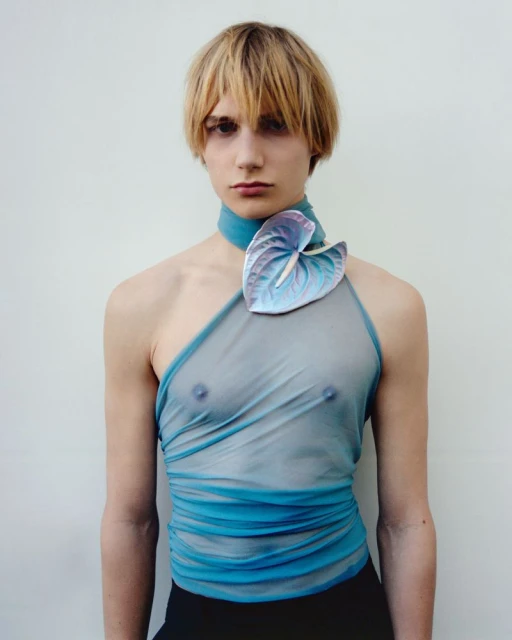 Person poses in a blue, sheer top with a large flower on the shoulder against a plain white background in a studio shot. Fashion forward.