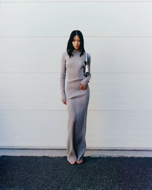 Adult female fashion model in a full-length ribbed knit dress with purse in an outdoor setting in front of a white wall and asphalt.