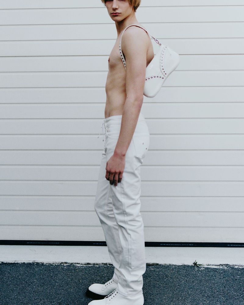 A slender, fair-skinned young adult poses in white jeans and shoes, accessorized with a white handbag, against a textured wall backdrop.