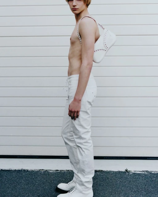 A slender, fair-skinned young adult poses in white jeans and shoes, accessorized with a white handbag, against a textured wall backdrop.