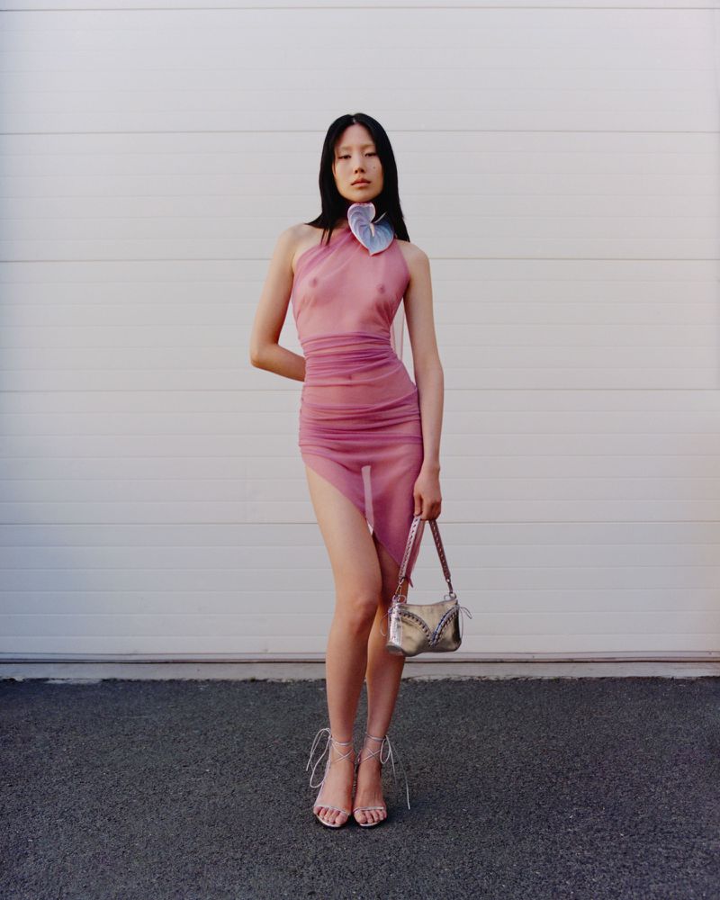 An Asian female model poses against a white wall wearing a pink dress and sandals while holding a silver purse. The scene is set outdoors.