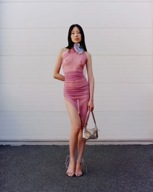 An Asian female model poses against a white wall wearing a pink dress and sandals while holding a silver purse. The scene is set outdoors.