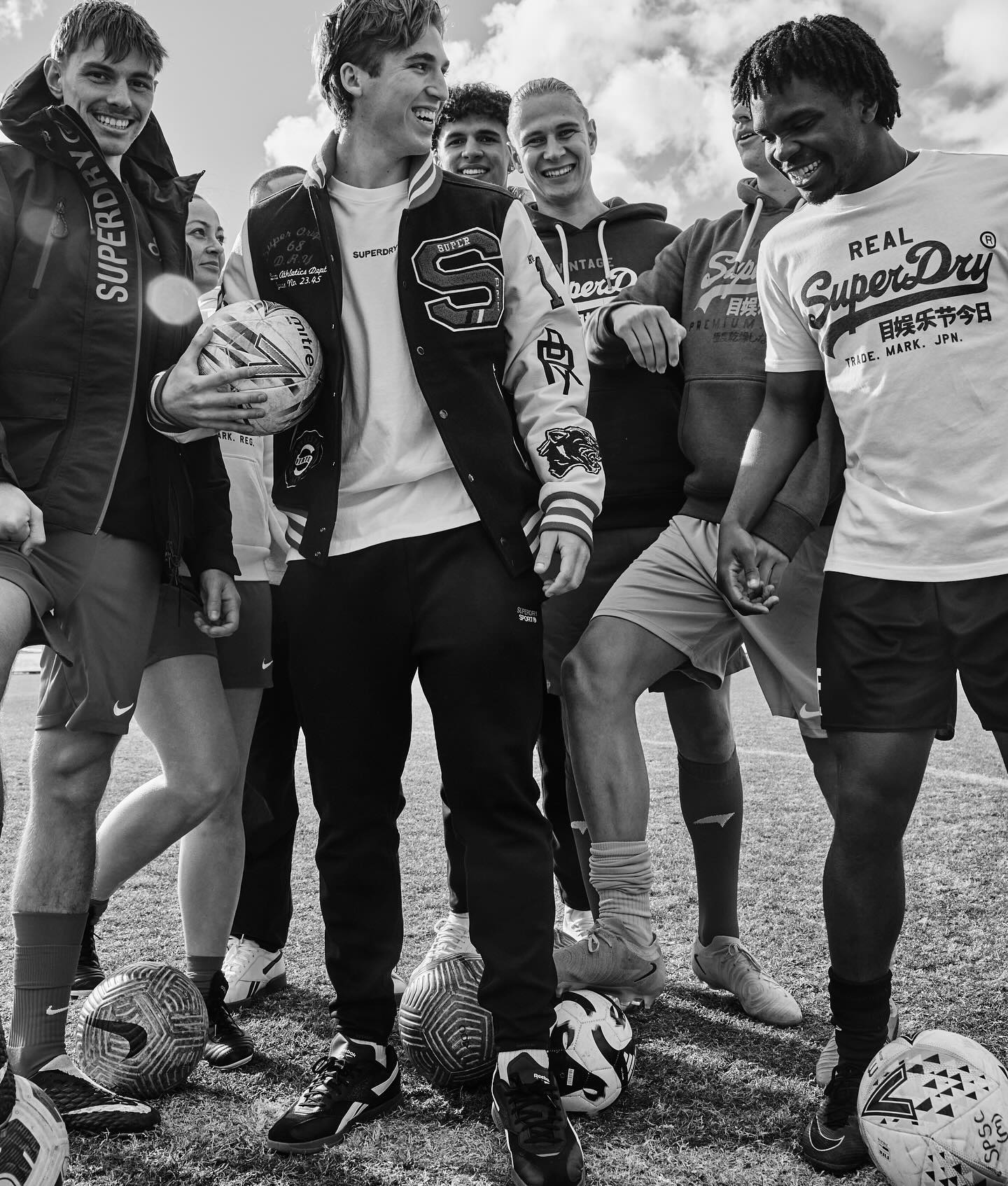 Monochrome photo of a diverse group of smiling people outdoors surrounded by soccer balls, wearing Superdry branded clothing.