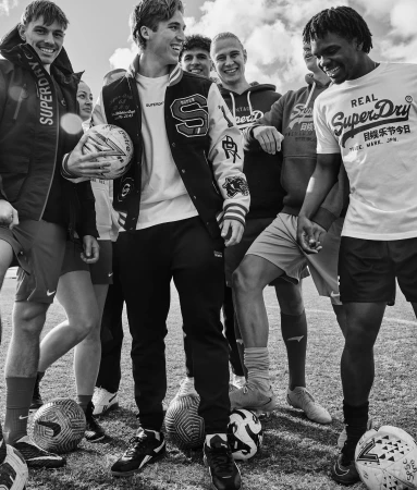 Monochrome photo of a diverse group of smiling people outdoors surrounded by soccer balls, wearing Superdry branded clothing.