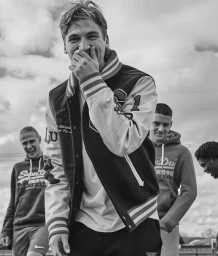 Young man laughing and covering mouth wearing a varsity jacket with three other young men smiling in the background outdoors.