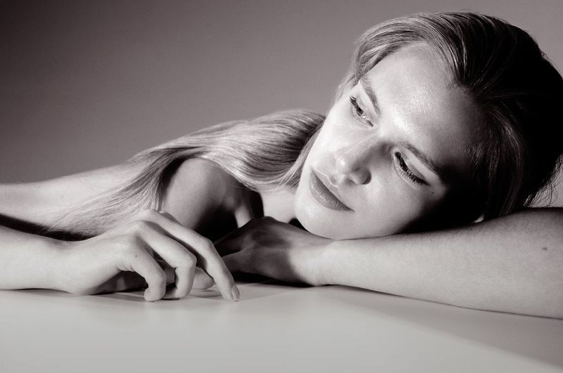 Monochrome portrait of a fair-skinned young woman with long blond hair resting her head on her arms, indoor studio lighting.
