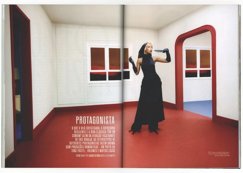 Editorial fashion shot of a woman in black gown with knife in an interior with red floor and trim, viewed through a magazine spread.