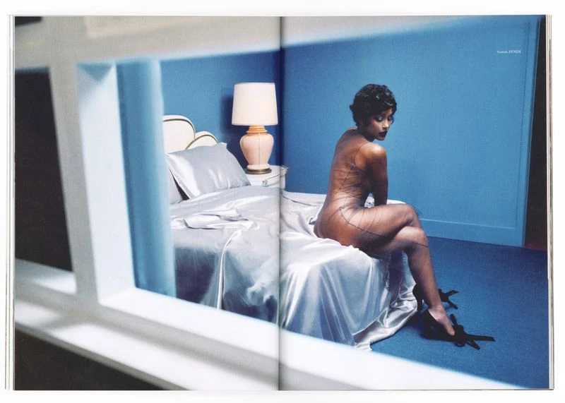 Woman sits on bed in a studio with blue walls, modeling sheer netting over her body while wearing black heels, next to a bedside table.