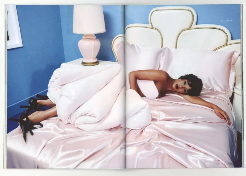 Woman poses on a pink satin bed with a white headboard, next to a white nightstand with a pink lamp against a blue wall indoors.