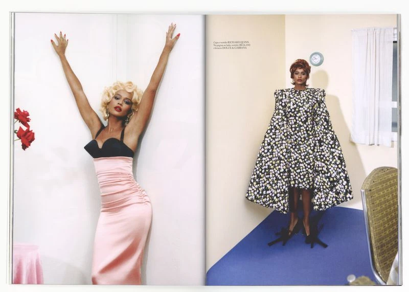 A woman in a pink gown and blonde wig poses next to a mannequin in a floral dress in a studio with white and blue walls.