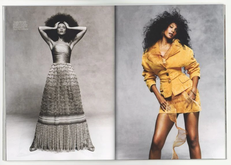 Editorial fashion spread with two women in gowns, one in a tan gown, the other in a golden suit, against a grey backdrop.