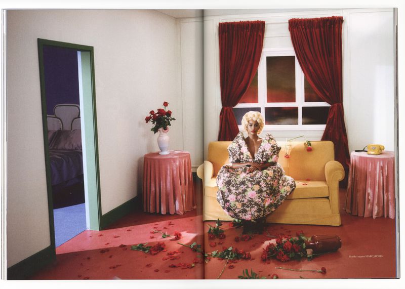 Blonde woman in floral dress sits on yellow couch amid scattered roses in a staged room with pink table and draped red curtains.