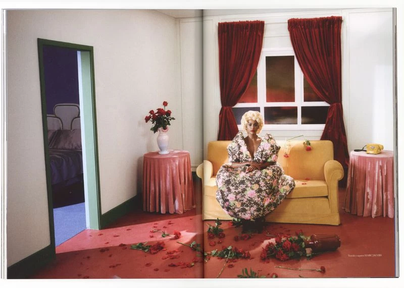 Blonde woman in floral dress sits on yellow couch amid scattered roses in a staged room with pink table and draped red curtains.