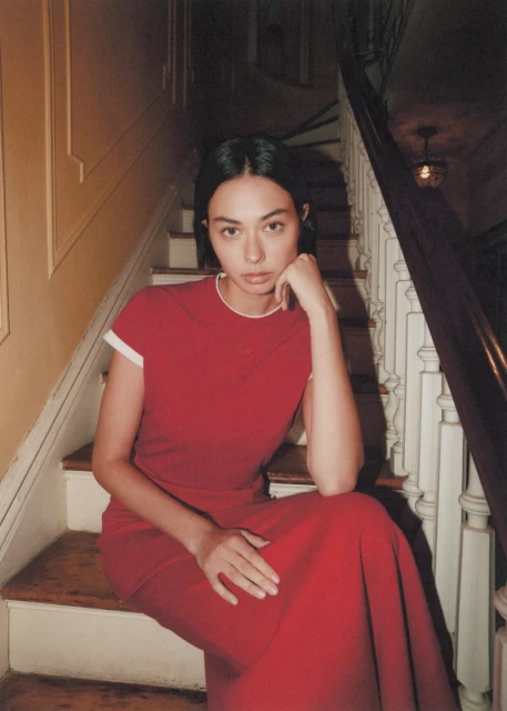Woman in a red dress sitting on wooden stairs indoors, looking at the camera with a neutral expression, hand on face.