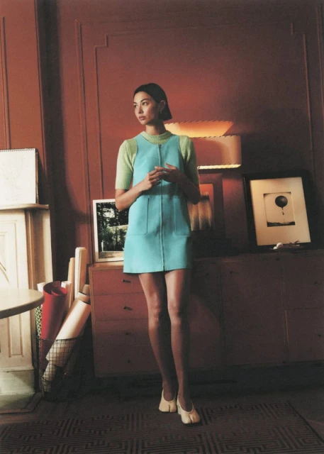 A woman poses in an indoor setting with light brown walls, wearing a green top and a short, light blue dress with white shoes.
