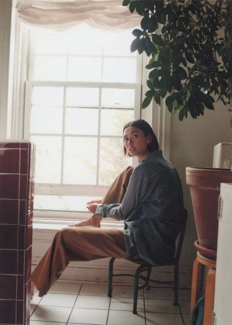 Young woman sits by a bright window in a patterned shirt and ochre pants with a plant and other objects in the background.