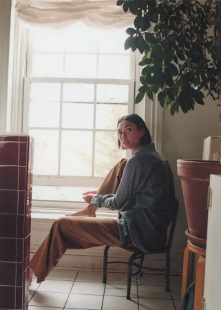 Young woman sits by a bright window in a patterned shirt and ochre pants with a plant and other objects in the background.