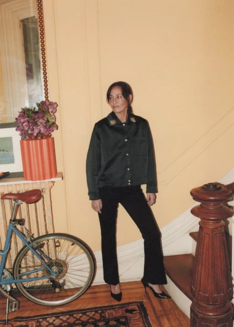 Woman in a black jacket and pants poses on stairs with a bicycle and vintage rug in an indoor environment with soft lighting.