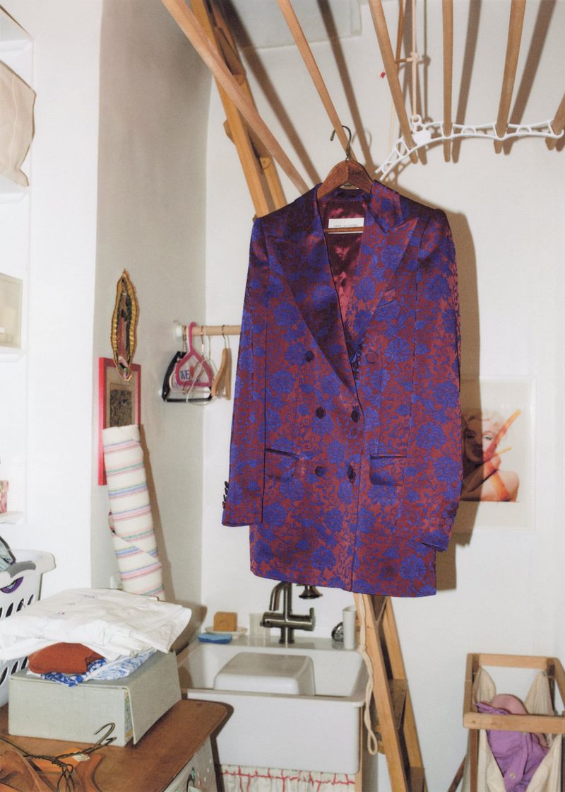 A maroon and purple floral blazer hangs in a bright, cluttered laundry room with a sink, drying rack, and various household items.