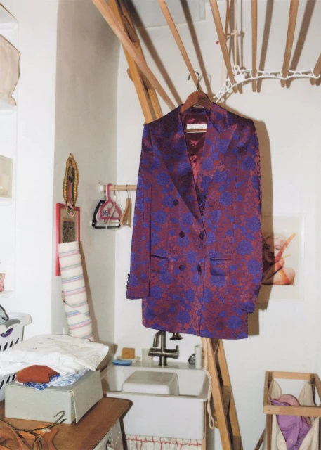 A maroon and purple floral blazer hangs in a bright, cluttered laundry room with a sink, drying rack, and various household items.