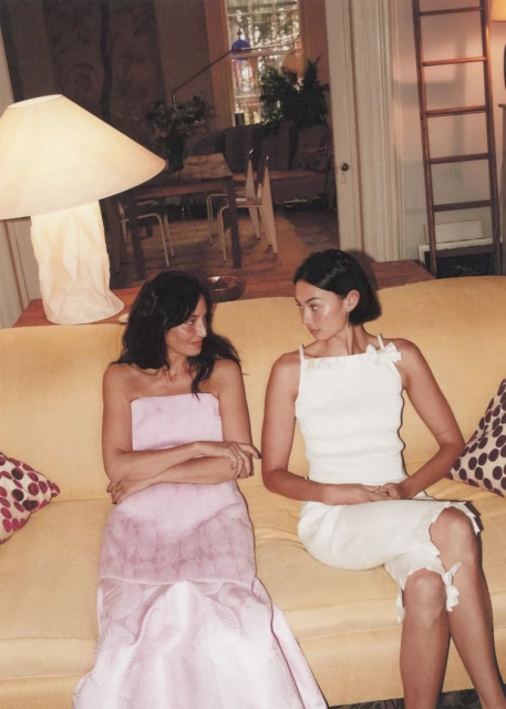 Two women in dresses sit on a yellow couch in an indoor setting. One woman wears a pink dress, the other a white dress.