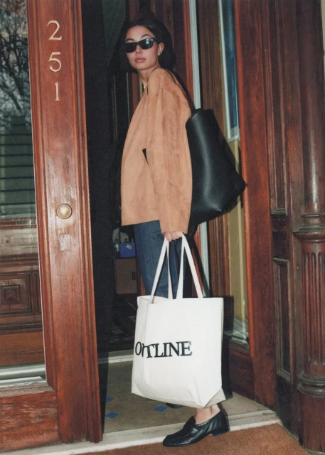 Woman in sunglasses and brown jacket, holding tote bag near building entrance with visible address, capturing a casual urban style.