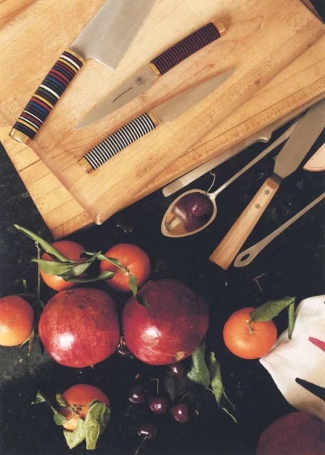 Culinary still life featuring knives on cutting board, and pomegranates, cherries, tangerines on a black countertop, lit with natural light.
