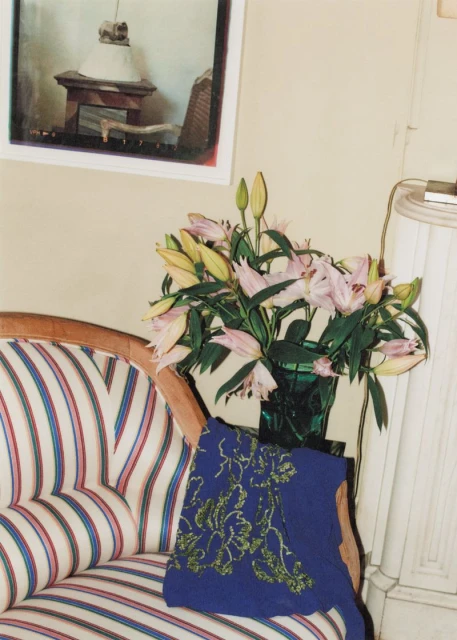A vintage-style interior features a striped settee with a blue embroidered throw pillow, a vase of lilies, and a framed image on the wall.