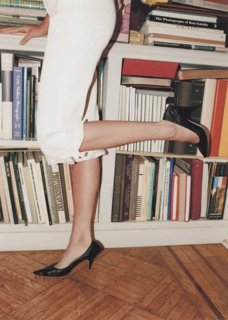 Woman poses with a black pump on a bookcase displaying books in a home. The woman wears white capri pants and is holding the bookcase.