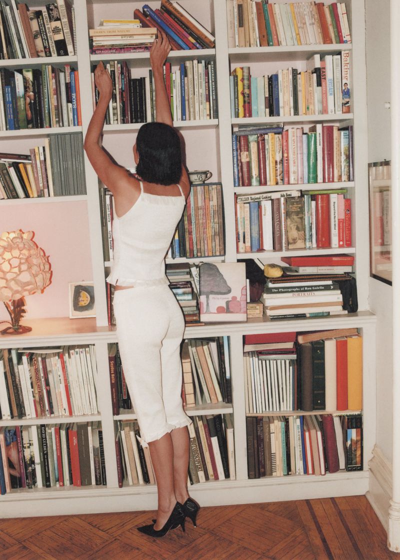 Woman in white outfit reaches for a book on a tall bookshelf in a home library with a hardwood floor and soft, diffused lighting.