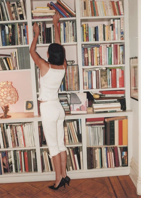 Woman in white outfit reaches for a book on a tall bookshelf in a home library with a hardwood floor and soft, diffused lighting.