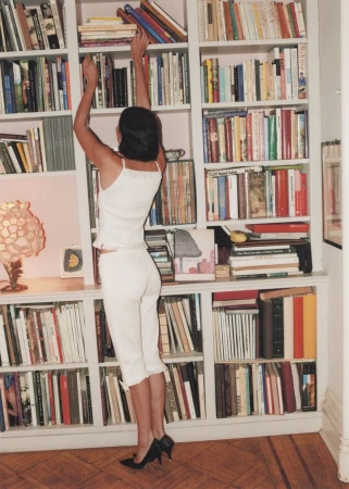 Woman in white outfit reaches for a book on a tall bookshelf in a home library with a hardwood floor and soft, diffused lighting.